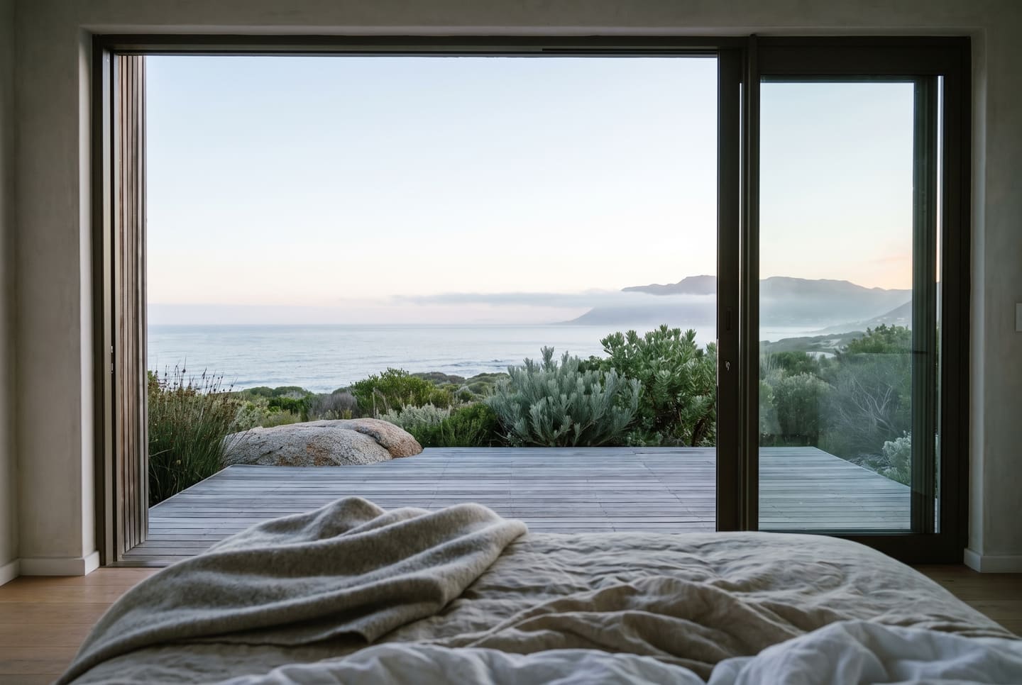 The view from a villa bed at first light: the glass wall open onto the deck, a granite boulder, fynbos and the sea under mist