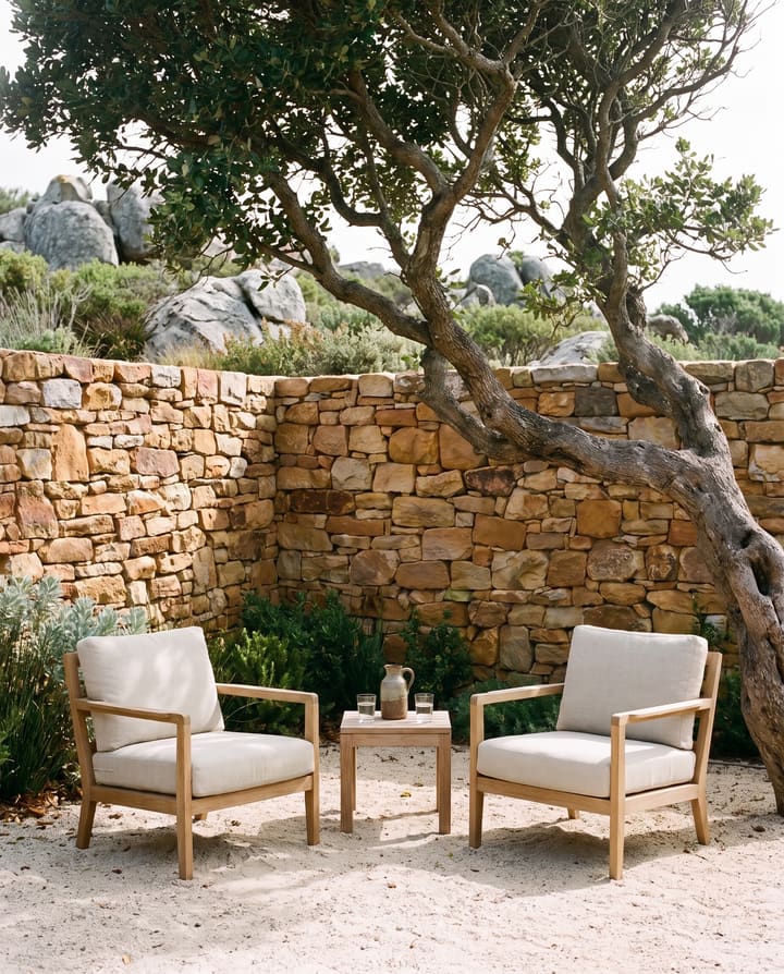 Two linen armchairs facing each other under a milkwood in a walled sandstone courtyard, a carafe of water on a low table