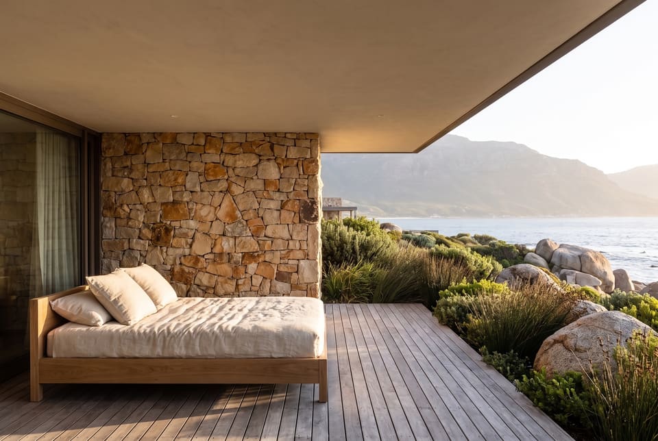 A linen day bed in the shade of a villa roof, late sun on the rubble wall, the mountain and the sea beyond