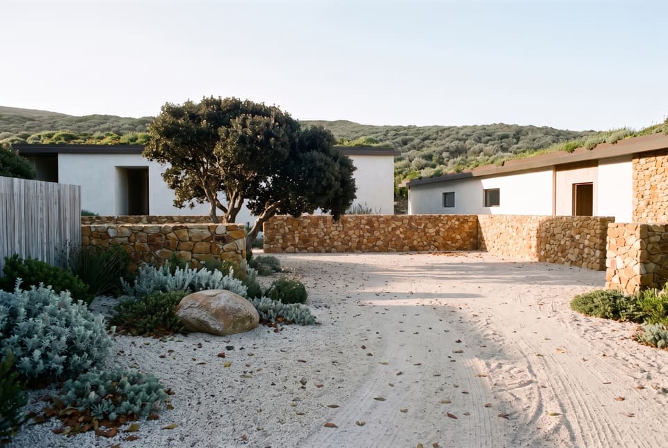 Early morning in the walled entrance court: raked pale gravel with fallen leaves, low rubble sandstone walls, one wind-bent milkwood, the limewashed therapy house and the long house beyond, a fynbos slope behind