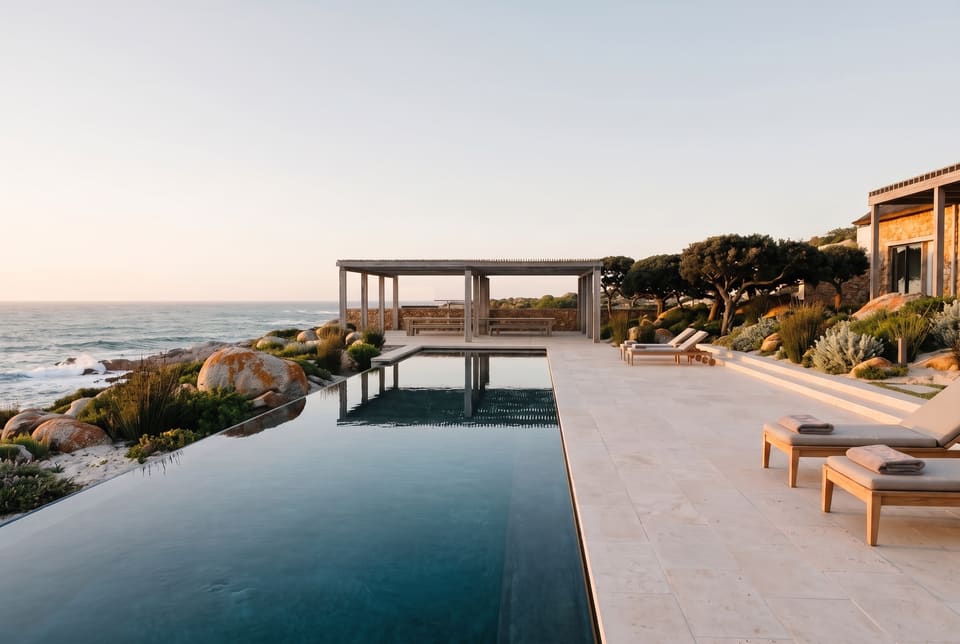 A long infinity pool on a limestone terrace in late sun, loungers beside it, the house and its pergola on the right, the open timber dining pavilion and milkwoods at the far end, the sea on the left
