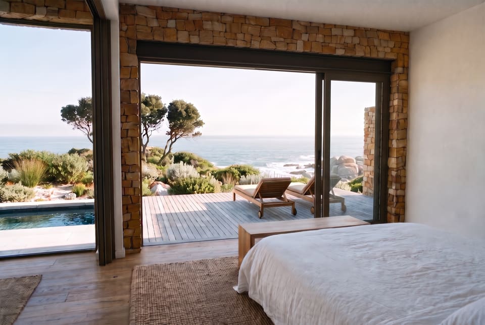 A villa bedroom with a white linen bed, a jute rug and oak floor, its glass wall slid open to a timber deck, a plunge pool, fynbos and the sea