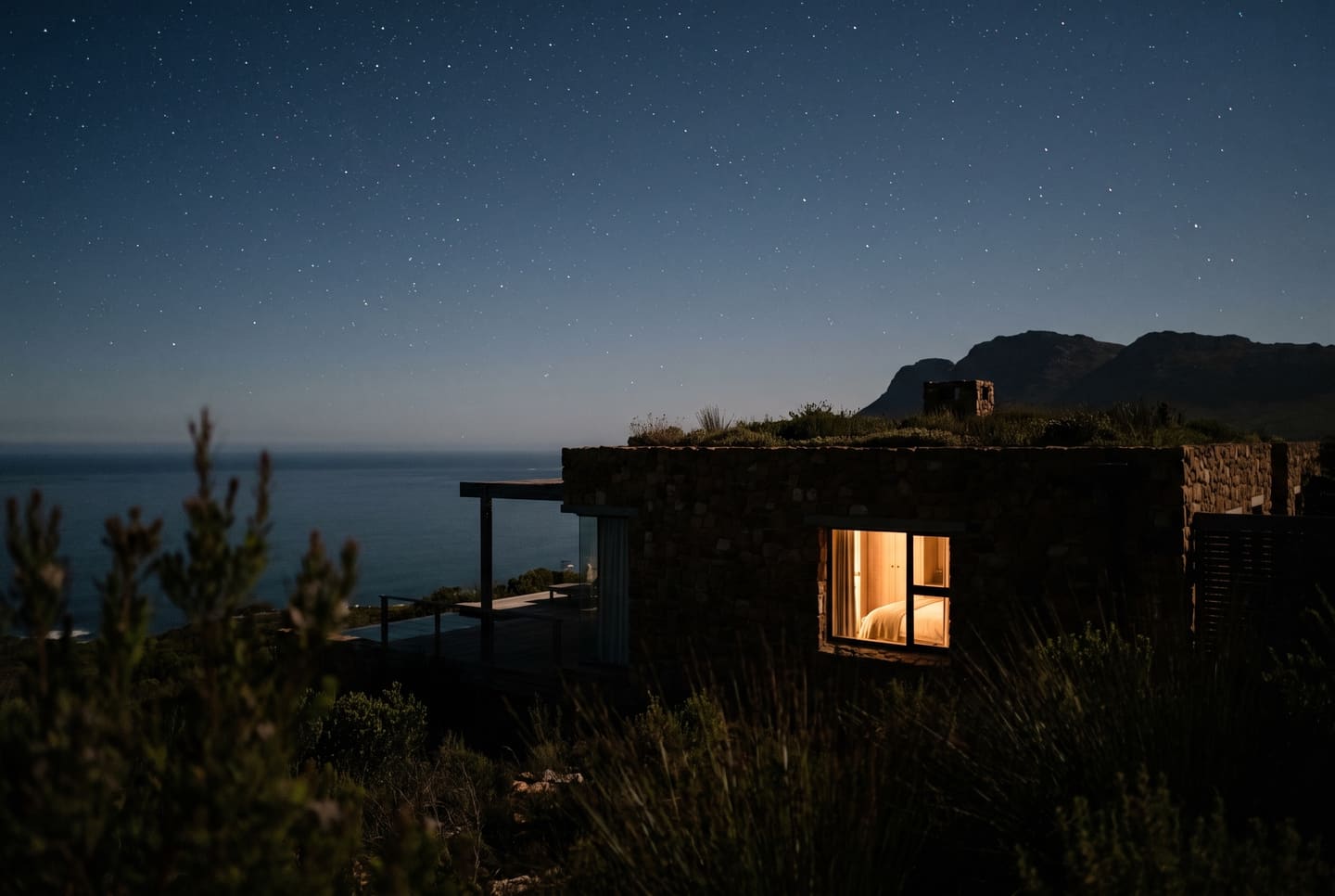 A guest villa at night under the stars, one window lit, the sea dark beyond