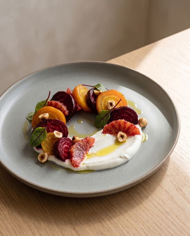 A plated starter: red and golden beetroot in a loose ring over labneh, with blood orange, toasted hazelnuts and beet leaves, on a grey stoneware plate