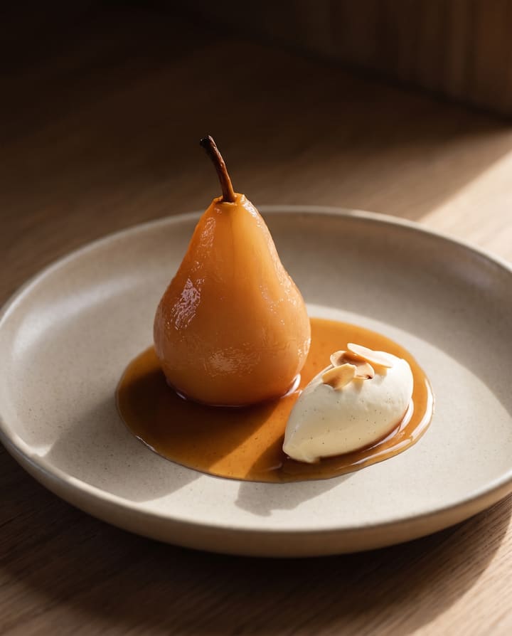 A pear poached in rooibos standing in its amber syrup, with vanilla cream and toasted almond flakes, on a stoneware plate