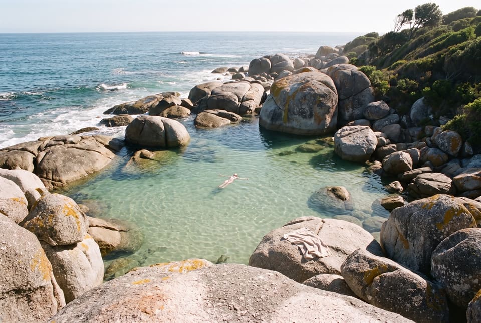 A clear rock pool among granite boulders at the sea’s edge, one swimmer floating, a towel on a boulder